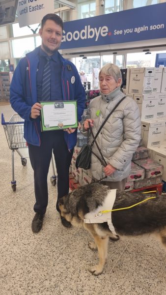 Gill Shepherd hands a Tesco's shop assistant a certificate for allowing her to collect donations at the store. Alba is smelling his feet.